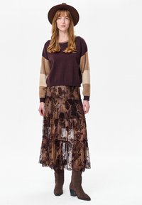 Bruin en beige gebreide trui met lange mouwen, gecombineerd met een gelaagde, doorschijnende maxirok met bloemenpatroon in aardetinten en bruine laarzen met hak.