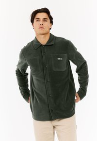 Muškarac u tamnozelenoj flis jakni na dugmad s dva džepa na prsima, stoji s rukama iza leđa na bijeloj pozadini.