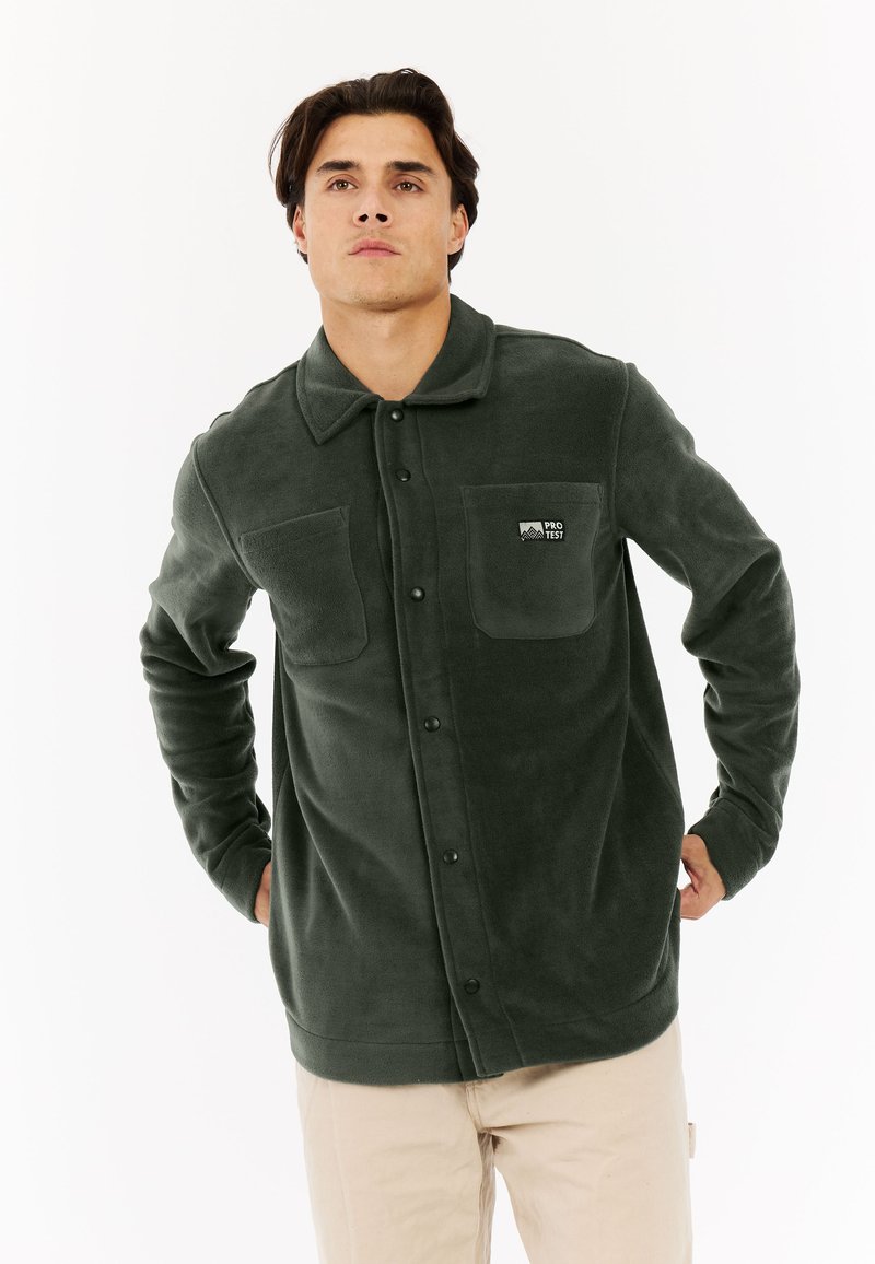 Muškarac u tamnozelenoj flis jakni na dugmad s dva džepa na prsima, stoji s rukama iza leđa na bijeloj pozadini.