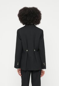 Person med krøllete hår iført en svart skreddersydd blazer med sølvknapper på baksiden og svarte bukser, stående med ryggen til.