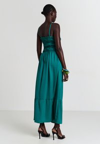 Eine Frau steht und trägt ein teal-farbenes, ärmelloses Maxikleid mit gerafftem Oberteil und schwarze hochhackige Sandalen. Sie hält Armbänder und Ringe in der rechten Hand.