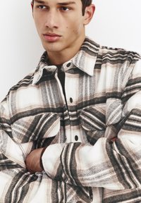 Camicia di flanella a quadri nei toni del bianco, marrone e nero; presenta chiusura con bottoni frontali, due tasche sul petto e una vestibilità comoda con tessuto strutturato.