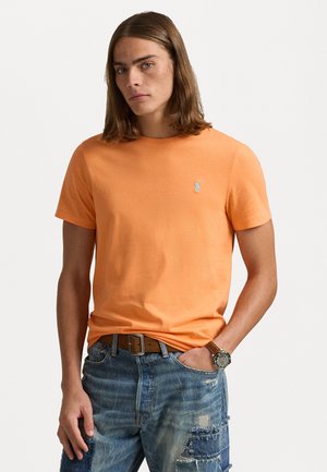 Oranje katoenen t-shirt met een ronde hals, korte mouwen en een klein blauw logo op de linkerborst. Gedragen met denim cargo shorts.