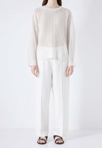 Pull en tricot blanc côtelé avec un col rond, un design à plusieurs couches, et des manches longues, associé à un pantalon blanc ample et des sandales noires.