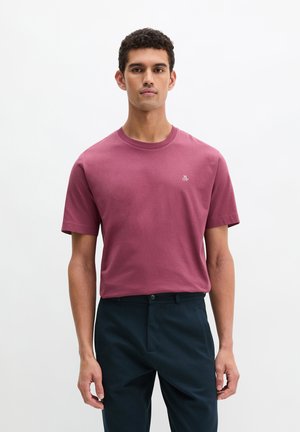 Giovane uomo in piedi, che indossa una maglietta viola chiaro a maniche corte con un piccolo logo e pantaloni blu scuro, su uno sfondo chiaro e uniforme.