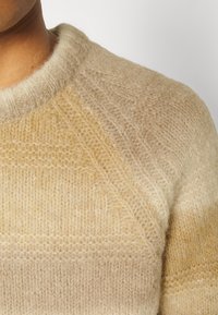 Beżowy sweter dzianinowy z okrągłym dekoltem, z teksturowanym wzorem i gradientowymi odcieniami od jasnego do ciemnego beżu.
