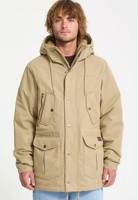 Beige waterproof jacket with a hood, multiple front pockets, zip closure, textured lining, and two snap buttons on the front.