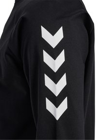 Maglietta nera a maniche lunghe con un motivo a zigzag bianco stampato verticalmente lungo la manica sinistra. Realizzata in un tessuto liscio e leggermente elastico.