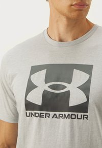 Siva kratka majica s kratkim rukavima koja sadrži veliki crni Under Armour logo s točkastim uzorkom, okrugli dekolte i opušteni kroj.