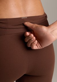 Barna sportleggings ribbeltartóval. Egy kéz kissé megfeszíti a derékpántot, megmutatva a puha, elasztikus szövet textúráját.