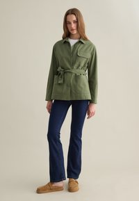 Giacca belted verde oliva con colletto e tasche frontali, abbinata a jeans svasati scuri e scarpe slip-on beige. Tessuto dalla consistenza liscia.