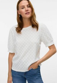 Bílý texturovaný top s krátkým rukávem a kulatým výstřihem, který má jemný vzorovaný pletený design. V kombinaci s modrými džíny.