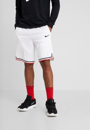 Uomo che indossa pantaloncini bianchi Nike con rifiniture a zigzag nere e rosse, calzini rossi, scarpe da ginnastica nere e una maglia a maniche lunghe nera con logo Nike.