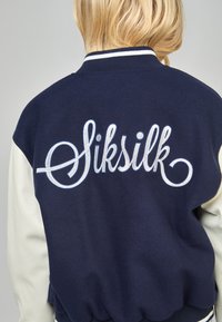 Mörkblå collegejacka med vita läderslackar. Har broderad "Siksilk" i ljusblå kursiv stil på baksidan. Ribbad krage.