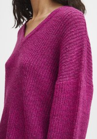 Pull en tricot fuchsia avec un col en V et une texture côtelée ; présente des épaules tombantes et de longues manches pour un ajustement décontracté.