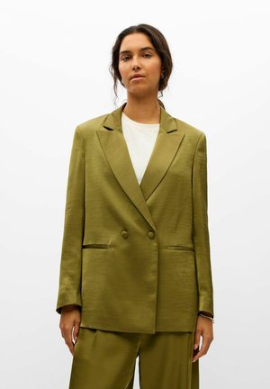 Une femme se tient devant un fond blanc uni, vêtue d’un tailleur croisé vert olive par-dessus un haut blanc.