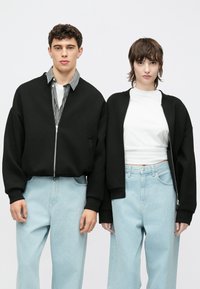 Zwarte jasjes met rits en wijde mouwen over witte en zwart gestreepte shirts. Lichtblauwe spijkerbroeken met een losse pasvorm, gedragen door twee modellen.