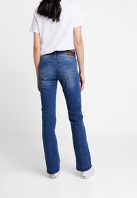 Femme debout, tournée de dos, portant un t-shirt blanc, un jean droit bleu et des baskets blanches sur un fond blanc uni.