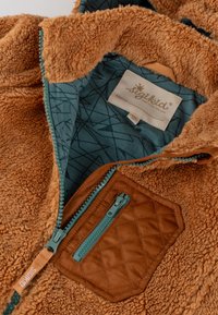 Braune Fleecejacke mit einer gesteppte sandfarbenen Tasche, türkisfarbener Reißverschluss und einem gemusterten türkisfarbenen Futter sowie einem Etikett mit dem Markennamen.