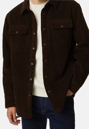 Brązowa kurtka koszulowa z sztruksu z dwoma kieszeniami na piersiach, zatrzaskami i luźnym fasonem. Nosi się ją na białym swetrze o fakturze oraz ciemnych dżinsach.