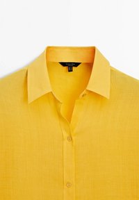 Chemise en lin jaune avec un col pointu, un bouton devant et une texture légère. Présente une petite étiquette à l'intérieur du col.