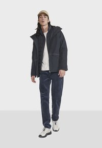 Doudoune noire avec capuche, zippers et poches latérales portée par-dessus une chemise claire. Un pantalon en velours côtelé bleu marine et des baskets blanches complètent le look.