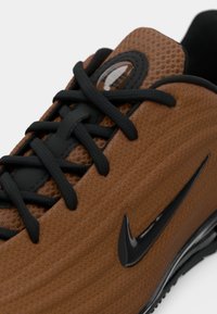 Καφέ υφασμάτινο αθλητικό παπούτσι με υφή στην επάνω επιφάνεια, μαύρα κορδόνια και γυαλιστερό μαύρο λογότυπο στο πλάι. Διαθέτει στρογγυλή τρύπα για τα κορδόνια και κομψό, αεροδυναμικό σχεδιασμό.