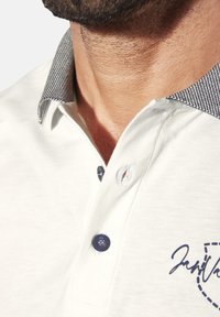 Kurzarm-Polohemd in Creme mit einem strukturierten Kragen in Marineblau und Weiß, ausgestattet mit einem Marineblauen Logo und zwei Knöpfen. Glattes Material.