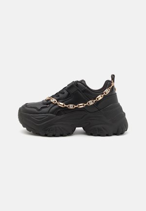 Czarny masywny sneaker z grubą podeszwą i dekoracyjnym złotym łańcuszkiem zdobionym cyrkoniami po boku na jednolitym tle.