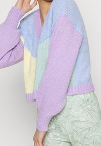Sweter dzianinowy z dekoltem w serek, oversize'owymi rękawami oraz wzorem w kolorach: lawenda, niebieski, żółty i miętowy. Włóczka o teksturowanej powierzchni.