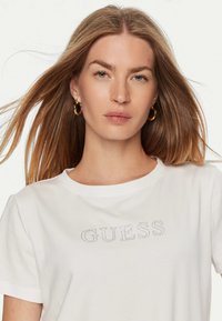 T-shirt bianco con la scritta "GUESS" in strass, scollo tondo e maniche corte, esposto su uno sfondo chiaro.