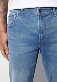 Jean en denim bleu clair avec une coupe droite, un design à cinq poches et un léger délavage, coutures contrastées et fermeture par bouton en métal.