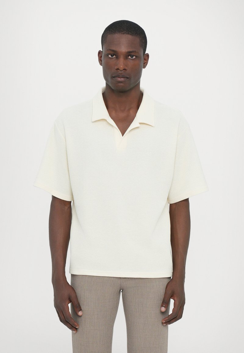 Ljusbeige kortärmad pikétröja med krage i en texturerad tygtyp. Har en v-ringning och en avslappnad passform. Matchas med grå slim-fit byxor.