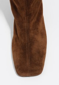 Brązowy zamszowy botek za kostkę z kwadratowym noskiem i widocznymi detalami szwów, ukazujący miękką fakturę i elegancki design.