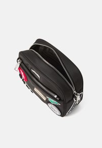 Borsa nera in nylon con due scomparti con zip, caratterizzata da vari patch, accessori in argento e una superficie liscia. Forma compatta e rettangolare.