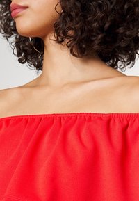 Haut rouge à épaules dénudées avec tissu froncé au niveau de l'encolure, en matière légère ; présente une texture lisse et une bordure élastique.