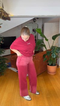 Červené sportovní tričko ve spojení s růžovými tepláky. Kalhoty mají boční kapsy a volný střih. Interiér místnosti zahrnuje rostliny a televizi.