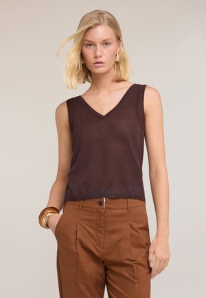 Blonde Frau trägt ein ärmelloses dunkelbraunes V-Ausschnitt-Top, hochgeschnittene braune Hose, goldene Creolen und Armreifen, eine Hand in der Tasche.