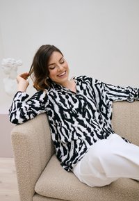 Femme souriante et détendue sur un canapé beige, portant un chemisier à motifs noir et blanc et un pantalon blanc, avec une sculpture de buste blanc en arrière-plan.