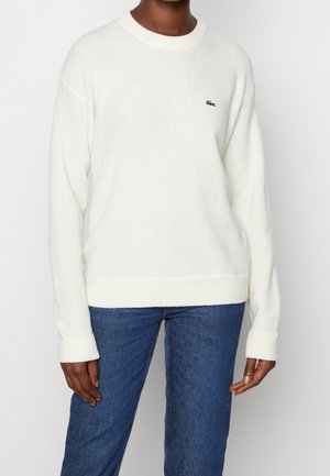 Osoba ubrana w biały sweter z długim rękawem z małym zielonym logo krokodyla na piersi oraz niebieskie dżinsy, stojąca na tle jednolitego tła.