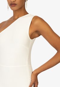 Witte geribde jurk met asymmetrische halslijn, aansluitende silhouet en een zijzak. De hand van het model rust op de heup.