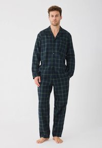Man standing barefoot wearing dark green and navy blue plaid long-sleeve pajama set with buttons, hands in pockets, neutral expression.