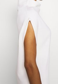 Weißer ärmelloser Rollkragenpullover mit seitlichen Schlitzen, weicher Stoff, lockere Passform, minimalistisches Design, glatte Textur und hoher Kragen.