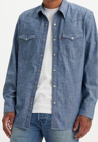 Camicia chambray blu scuro con bottoni a pressione, due tasche sul petto, design con yoke appuntito, maniche lunghe e texture sottile. Indossata sopra una t-shirt bianca.
