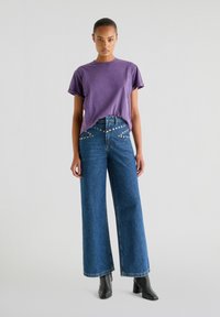 Lilla bomulds T-shirt og vide blue denim bukser med metalnitter langs taljen, præsenteret på en neutral baggrund.