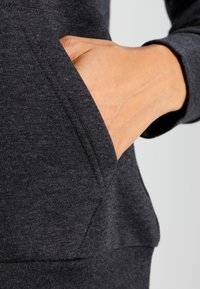 Donkergrijs sweatshirtstof met een kangoeroezak, met een hand die erin rust. Het materiaal lijkt zacht met een gladde textuur.