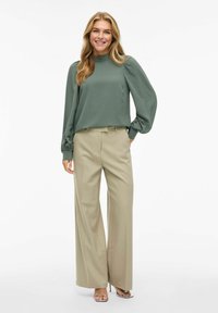 Blouse verte à manches longues avec un col rond et des manches froncées, associée à un pantalon beige taille haute à jambes larges et des talons à lanières.