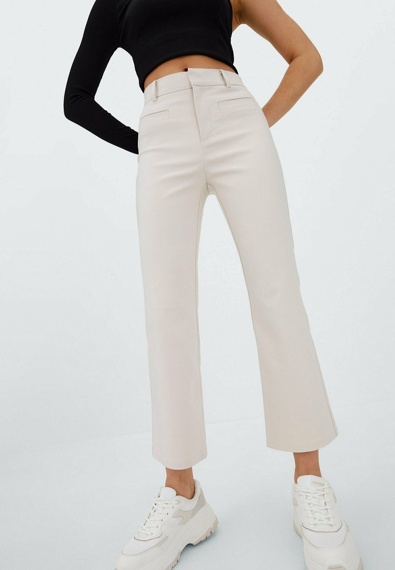 High-waisted, cream-colored, flared pants made of smooth fabric; features two front pockets and tailored seams, paired with white sneakers.