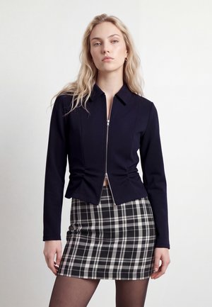 Une femme blonde présente une veste bleu marine zippée sur le devant et une jupe à carreaux noirs et blancs sur un fond clair uni.