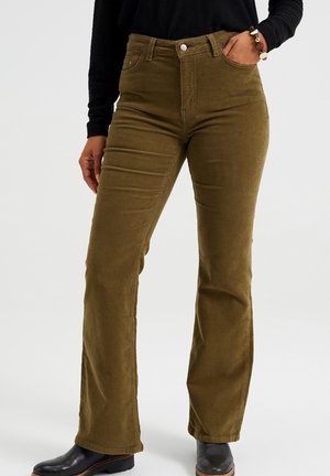 Vrouw draagt olijfgroene corduroy hoge taille flared broek met zwarte laarzen en een zwart top met lange mouwen, één hand in de zak.